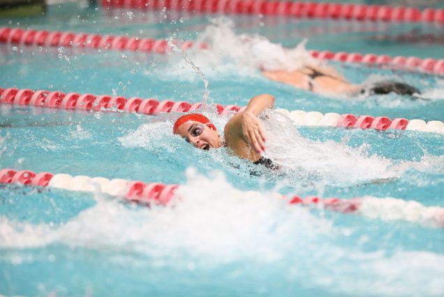 Photo of Julimar Avila (Sargent’19) during a swim meet at BU; in the photo she swims between lanes lined with red and white plastic lines and wears a red swim cap and googles.
