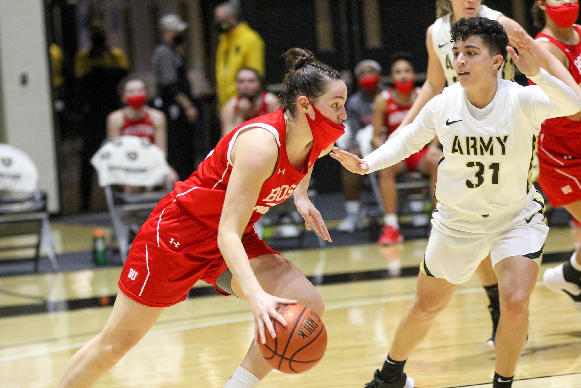 Photo of Riley Childs (COM’22) dribbling a basketball game during a game against the team “ARMY” in white jerseys, a defender stands with their hands up behind her to her left. Childs wears a red face mask.