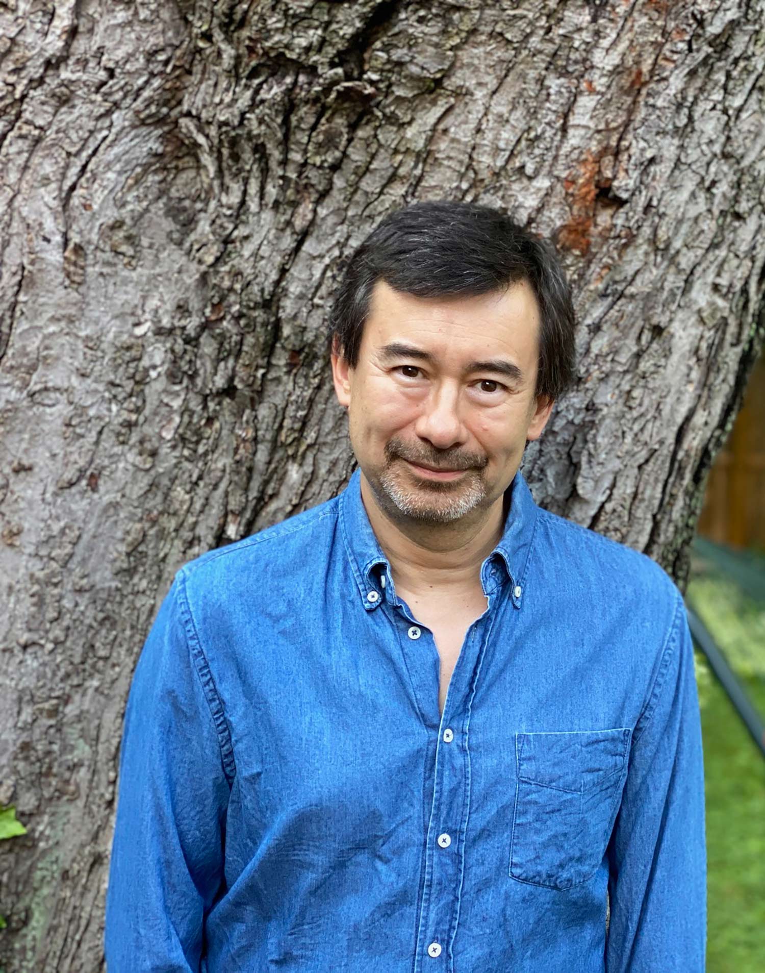 Portrait of Peter Ho Davies in a jean button down shirt in front of a tree trunk.