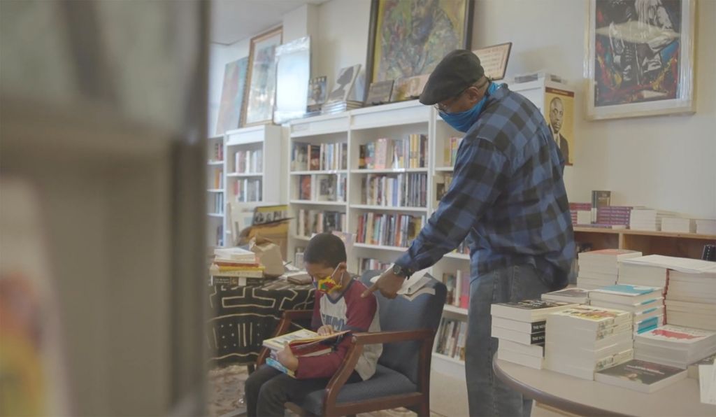 Frugal Bookstore owner Leonard Egerton points at a page as a young patron who sits in a chair in front of him reads a book. Both wear face masks.