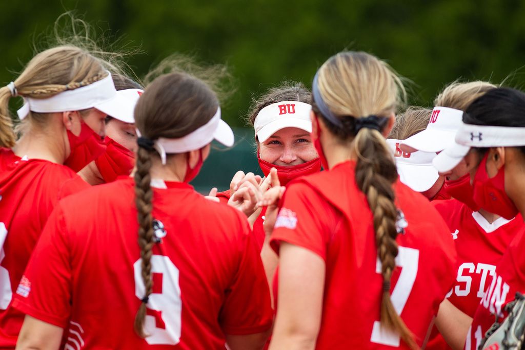 Photo of the Nicole Amodio (9) (Sargent’22) and the women’s softball team in a huddle, with red jerseys on and white visors.