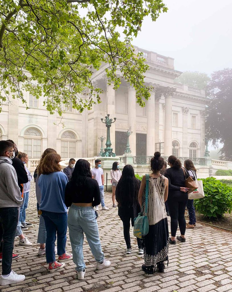 Photo of a group of students listening as they go on a tour of the Marble House in Newport. The students are outside on a stone path way and face the front of the opulent house on a foggy day.
