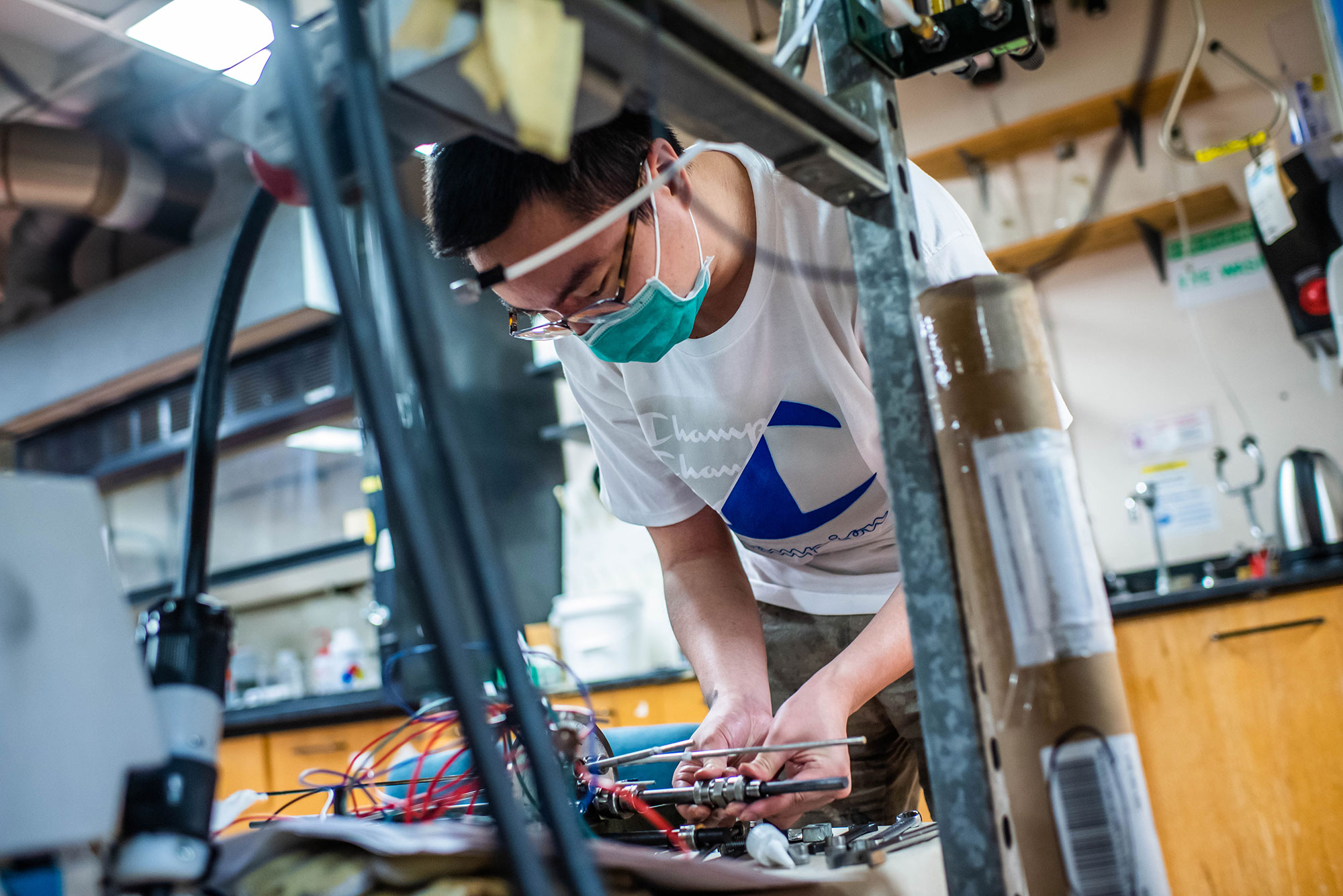 Kyle (Haoxuan) Yan (ENG’23) sets up an experiment to measure resistance on June 19, 2021. He wears glasses and a blue face mask and works with some wires. The shot is taken from slightly below with some machinery in the foreground.