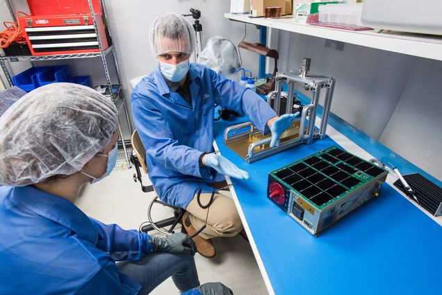 Photo of Emil Atz, ENG PhD Candidate ’22, gesturing with his hands and talking with Jacqueline Bachrach (ENG’23), seen at left in profile, about the CuPID (Cusp Plasma Imaging Detector) June 30 in the Walsh Lab in Photonics. Both are on the team which built the spacecraft, but Bachrach, who designed software for CuPID remotely, was seeing it for the first time. Both wear blue lab coats, hair bonnets, face masks and gloves. The device is about the size of a small brief case and has what look like mini-solar panels on top of it.