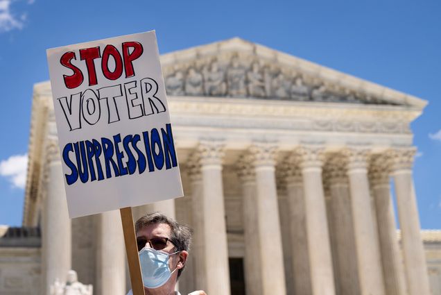 Photo of an activist at a rally against Sen. Mitch McConnell (R-KY) and Sen. Joe Manchin (D-WV), calling on them to eliminate the legislative filibuster and pass the “For The People” voting rights bill, outside the Supreme Court on June 23, 2021 in Washington, DC. The event was organized by a group called the Poor Peoples Campaign. The man wears sunglasses and a blue face mask and holds a sign that says “STOP VOTER SUPPRESSION” written in red, white and blue.