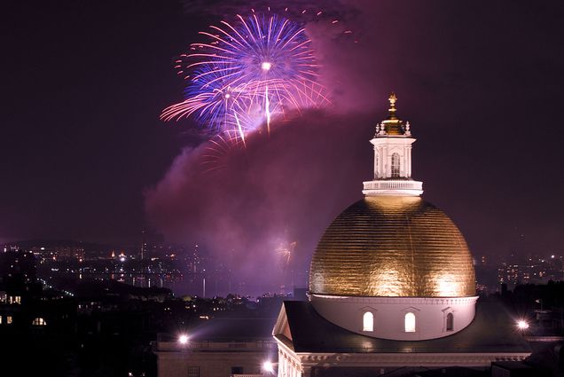 Photo of purple and pink fireworks exploding over the Charles River with the golden dome of the state house in the foreground at right.