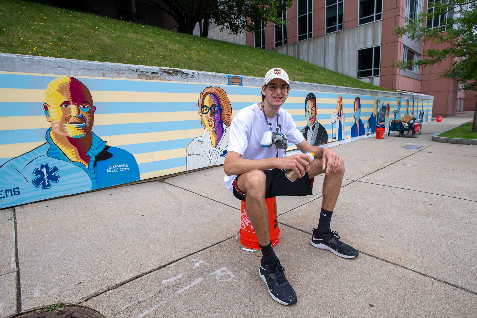 Photo of Sam Weinberger sitting on top of an orange home depot bucket, wearing white hat and t-shirt and shorts, in front of his recently painted mural at at St. Elizabeth Medical Center, which features colorful portraits of staff.