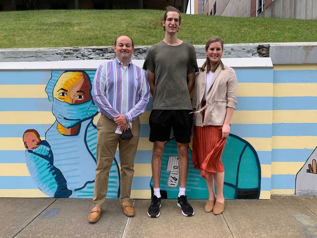 Photo of Aidan McDonough, in slacks and a purple striped shirt, Kelly Katapodis, in an orange skirt and tan sweater, and Sam Weinberger (CFA’21), in the center, in a greenish gray t-shirt, standing in front of the mural.