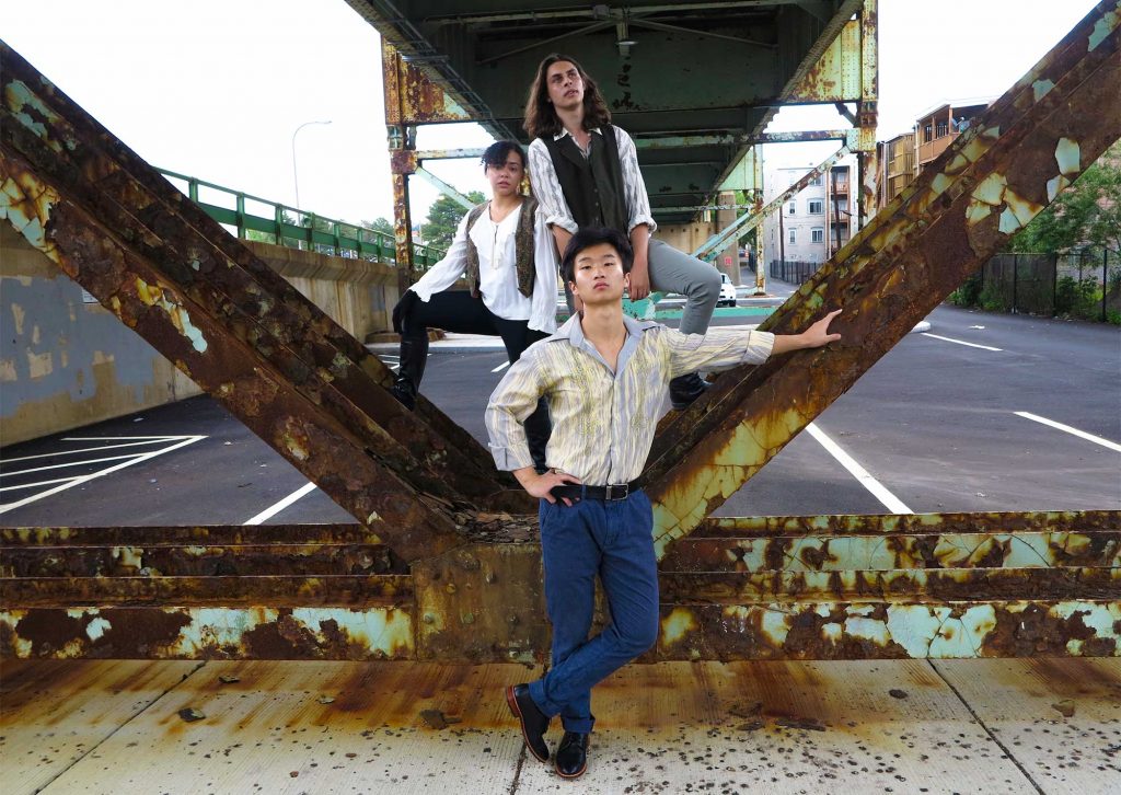 Photo of three young people dressed in button down shirts standing underneath a rusted overpass.