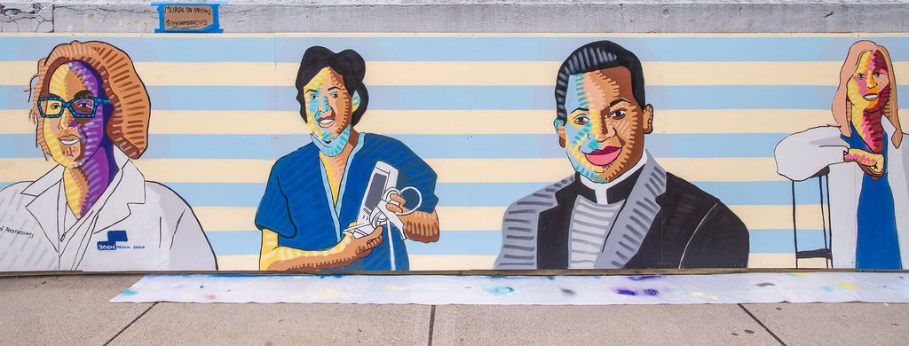 Photo of a 100-foot-long stretch of wall in front of St. Elizabeth Medical Center where Sam installed his mural. The mural has yellow and light blue stripes in the background, and vibrantly colored portraits of four hospital workers, include a doctor, a nurse holding a thermometer, a chaplain, and another doctor sitting in a chair. 