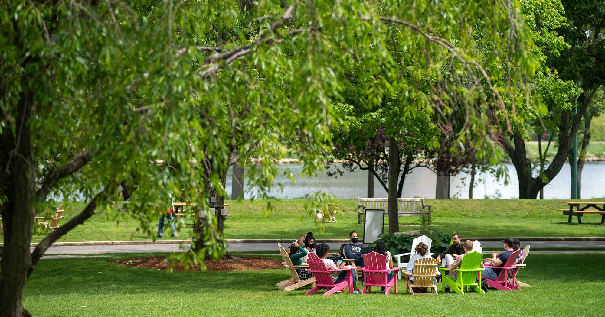 A class is lead in discussion sitting in a circle of colorful adirondack chairs beneath a tree on BU Beach, Boston University Charles River Campus.