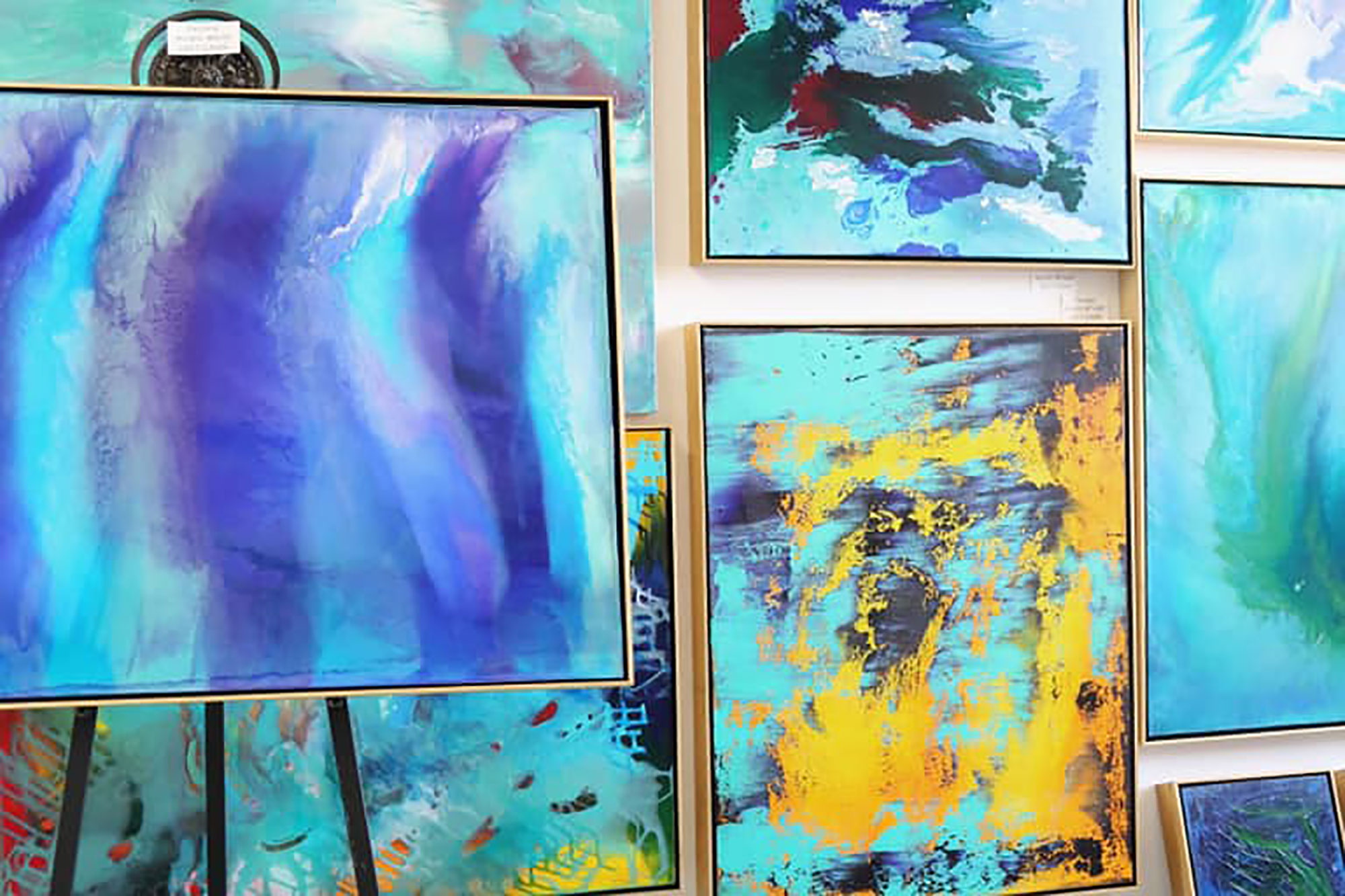 Photo of a wall of canvases painted in organic strokes of blue, teal, and gold, with an easel in the front left.