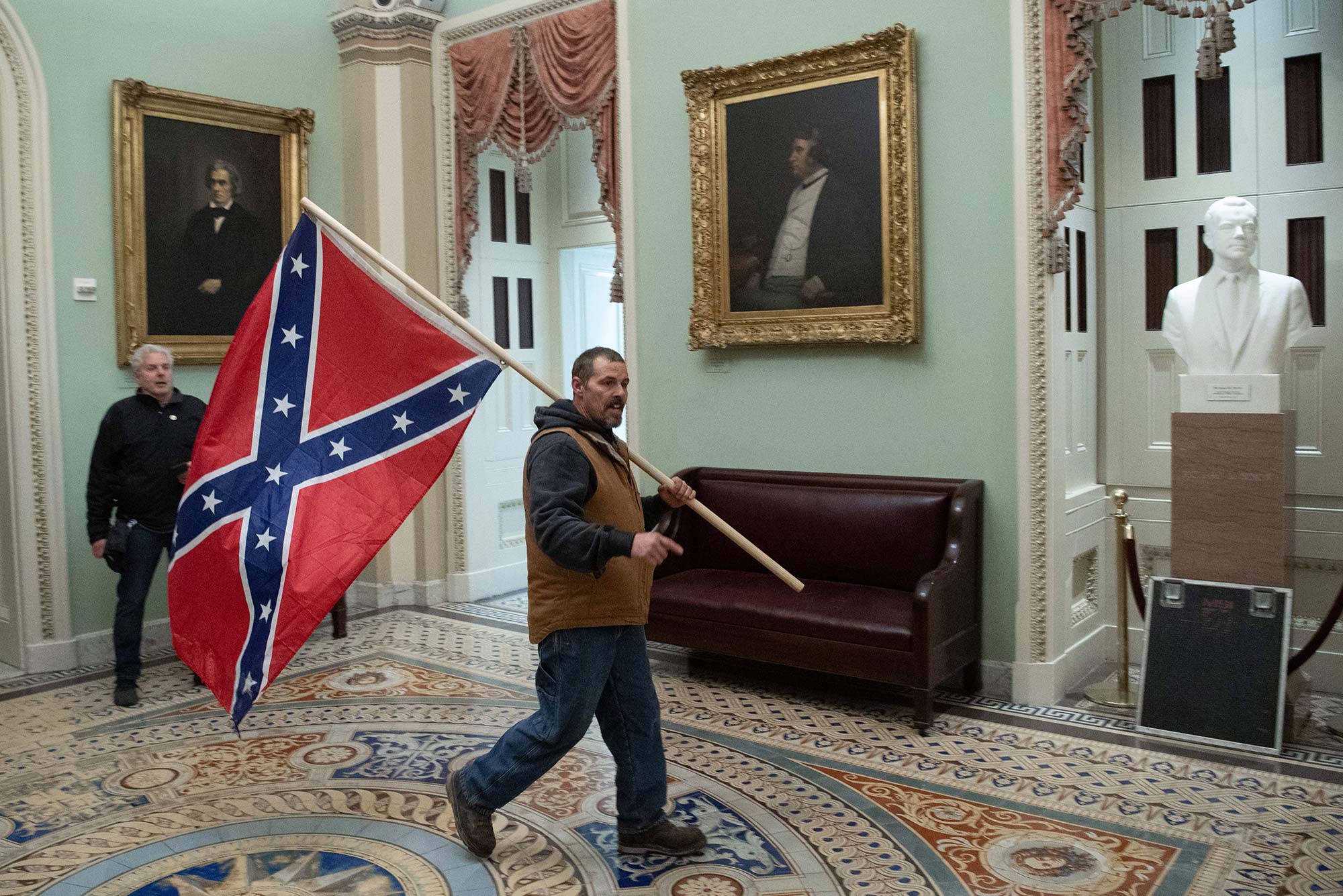 Photo of a supporter of US President Donald Trump, dressing in jeans, a tan vest and sweatshirt, carrying a Confederate flag in the US Capitol Rotunda during the insurrection on January 6, 2021, in Washington, DC. Behind him, a security guard looks on.