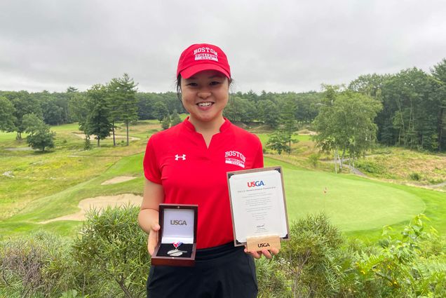 Photo of Hanako Kawasaki (Questrom’22) holding a USGA medal and plaque with her red Boston University polo and hat on. Behind her a golf course is seen on an overcast day.