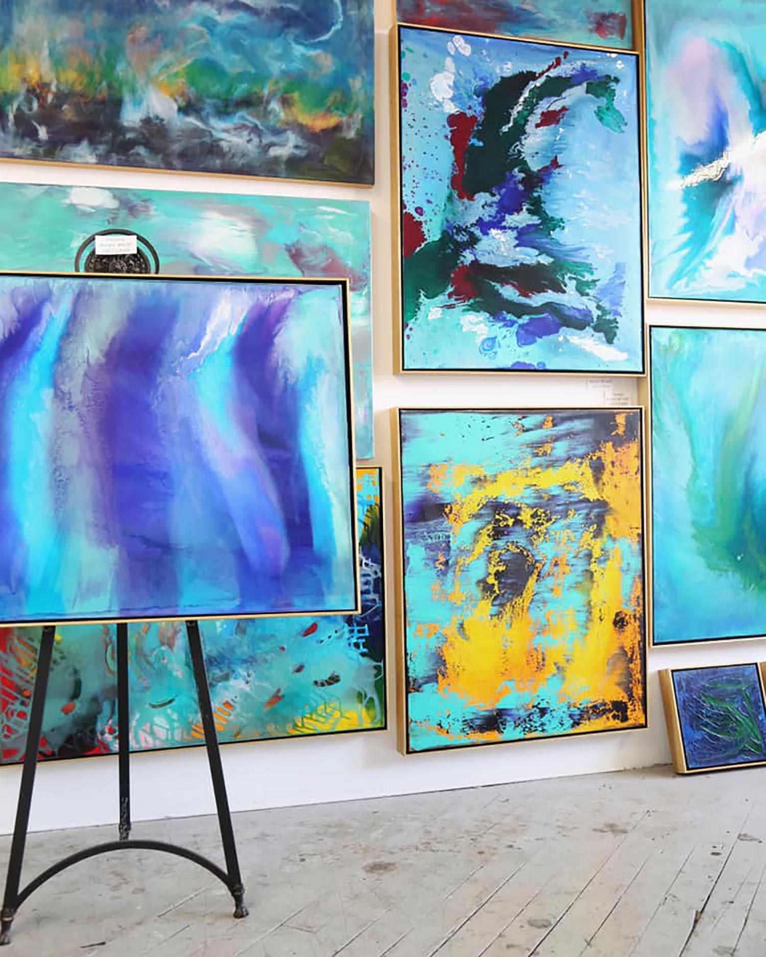 Photo of a wall of canvases painted in organic strokes of blue, teal, and gold, with an easel in the front left.