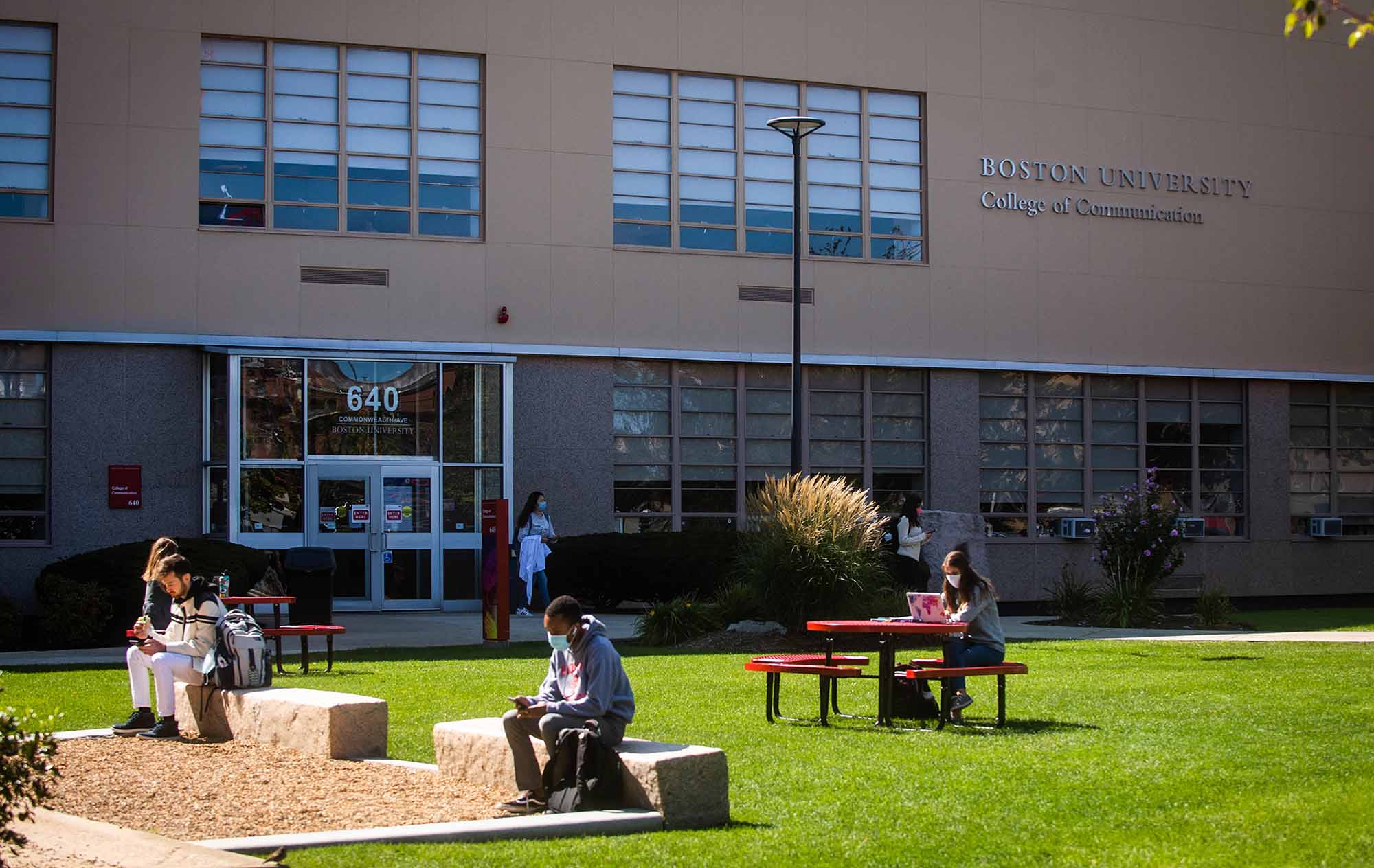 Students wearing protective face coverings sit at tables and benches on the lawn outside the Boston University College of Communication