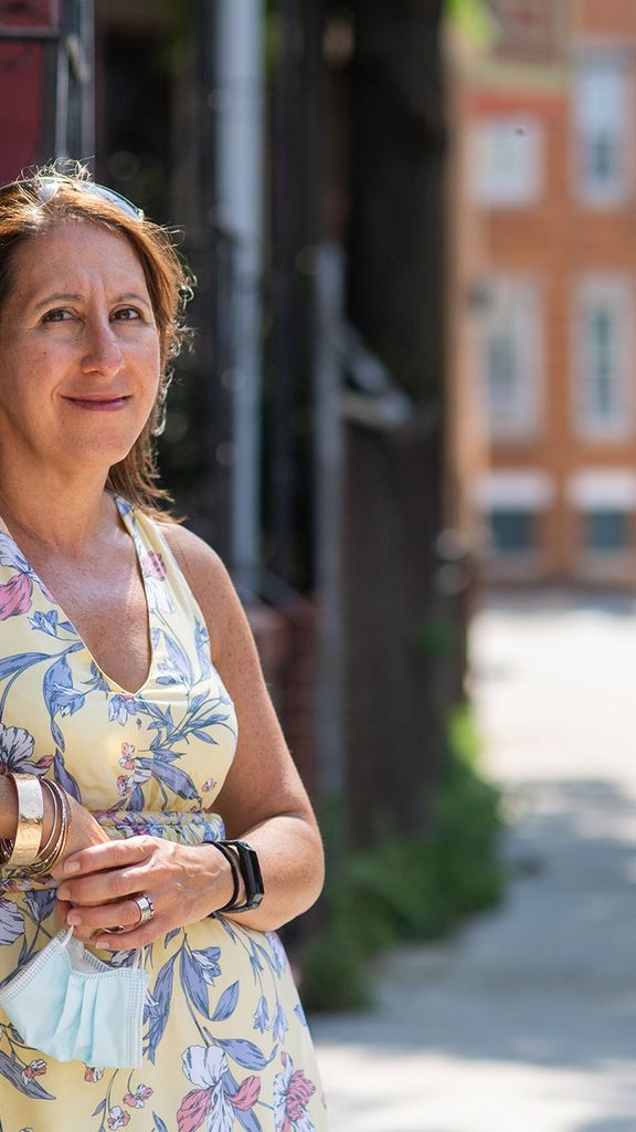 Photo of Roseann Bongiovanni (CAS’99, SPH’01), wearing a yellow sundress and leaning against a short wall outside of a building, holding her face mask in her hand, and smiling. Behind her, a person is seen carrying bags down the street.