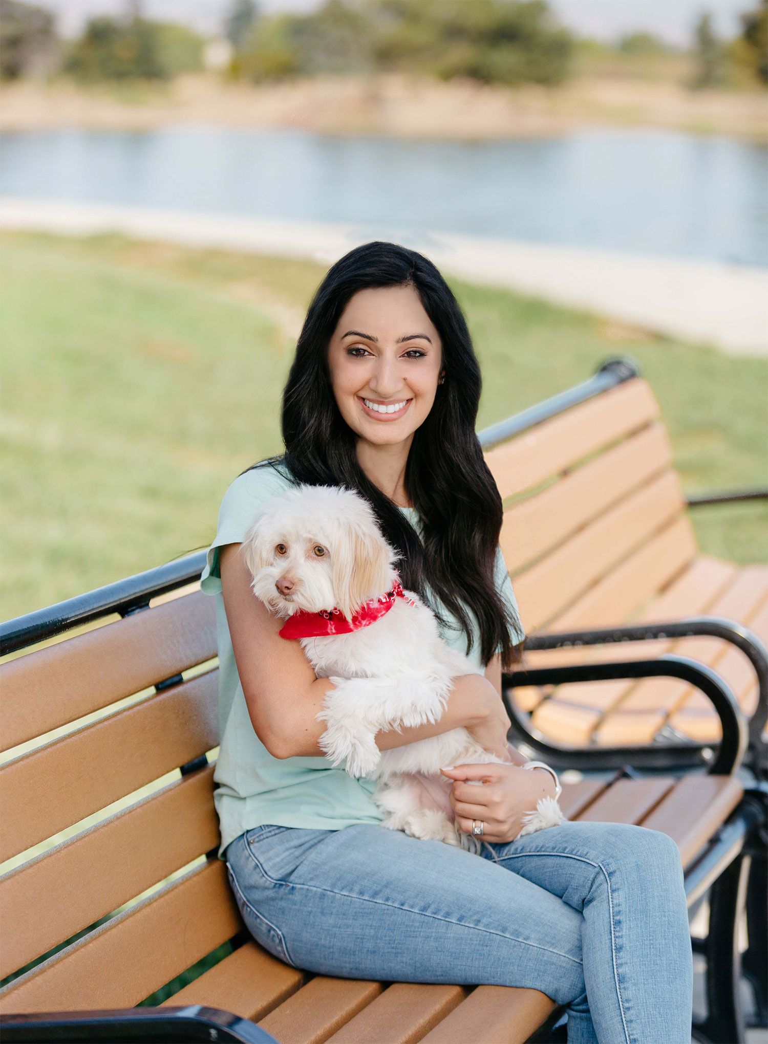 Photo of Rashi Khanna Wiese (CAS’06), who has long black hair, sits on a bench outside and smiles, with a small dog with a red bandana in her arms. Behind her, green space and a body of water is seen.