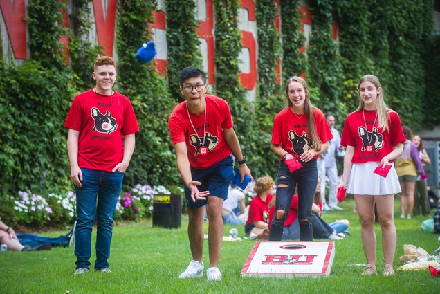 Photo of (Left to right) Joe D’Innocenzo (ENG 25) Derek Dong (QST 25), Kaylee Sanderson (QST 25) and Maddie Beery (QST 25) playing cornhole at the Terrier Tailgate on Sunday, August 29, 2021. Dong throws a blue sand bag towards the opposing whole. The students wear red shirts with terriers on them.