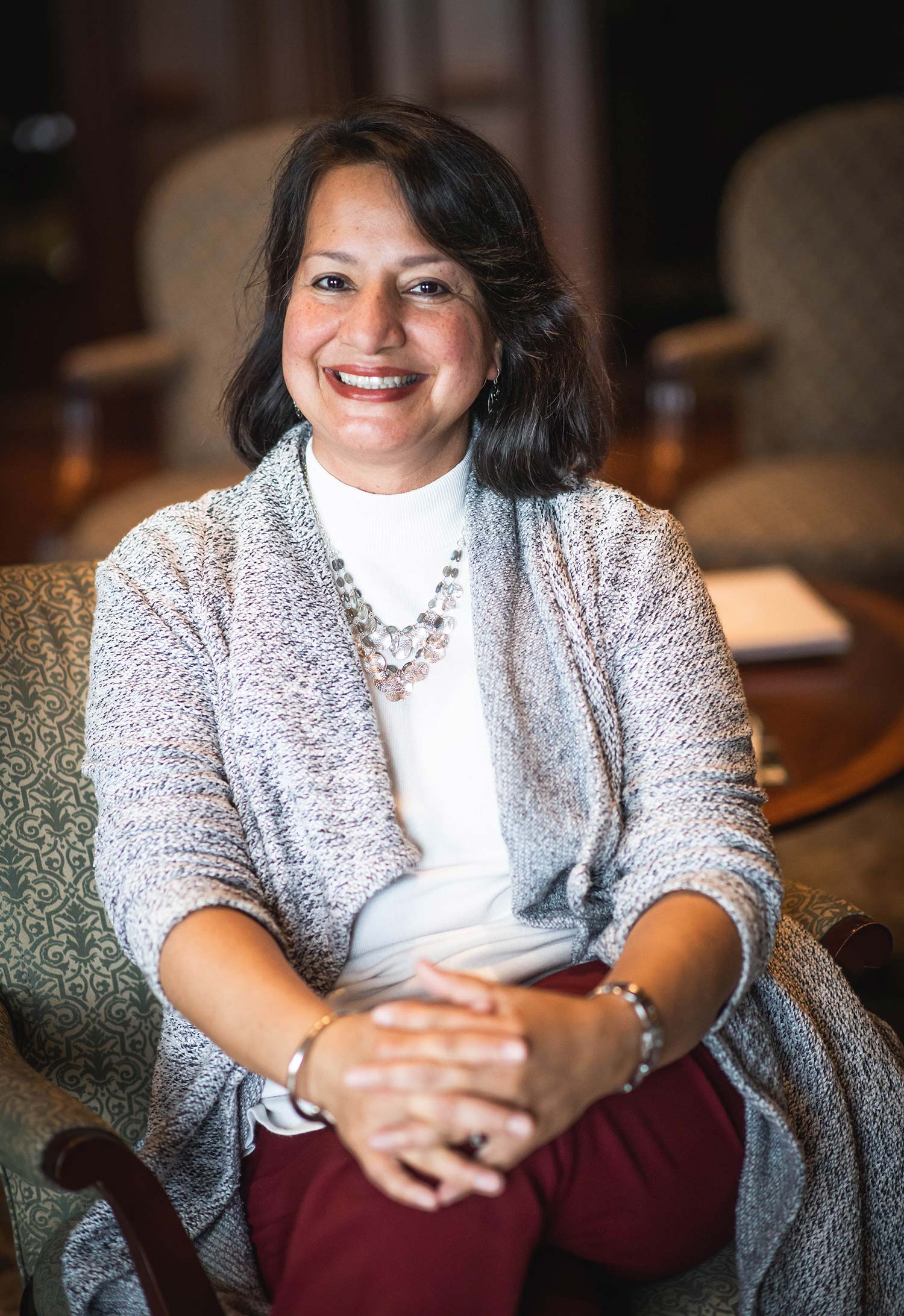 Ines Garrant with a gray sweater and white turtle neck on, sitting in an armchair with her hands clapsed at her knee. A dark wooden-paneled office space is seen behind her.