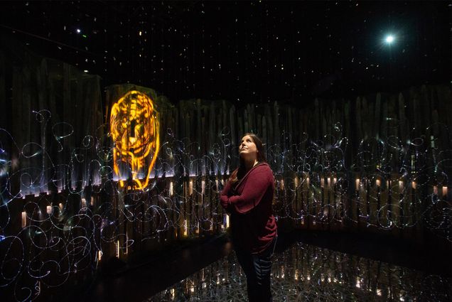 Photo of Kayleigha Zawacki (CFA’14,MFA’21) stands in the middle of her installation "Panthera tigris,” a 360 degree immersive mixed media experience on view at the Joan and Edgar Booth Theatre. Zawacki crosses her arms across her chest, and wears a crimson sweater. Around her are swirling lights and a neon figure of a tiger.