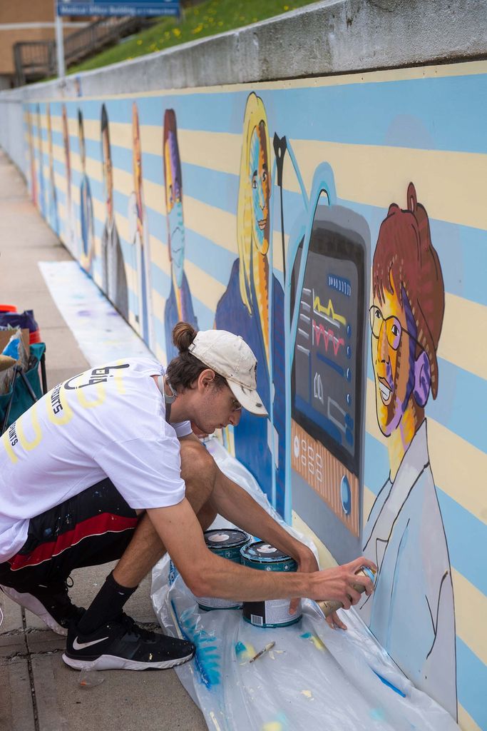 Photo taken from the side of Sam Weinberger as he uses spray paint to finish a portrait of a frontline healthcare worker next to a vitals monitoring machine. He crouches down by the roughly 3 foot wall outside of St. Elizabeth where the mural is being installed.