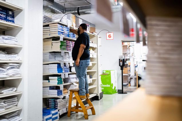 Photo of Terrence Samuel organizing textbooks at the BU Bookstore on August 18, 2021. He wears jeans and a black t-shirt, and stands on a wooden stool. In the foreground, the labels on a bookshelf are seen.