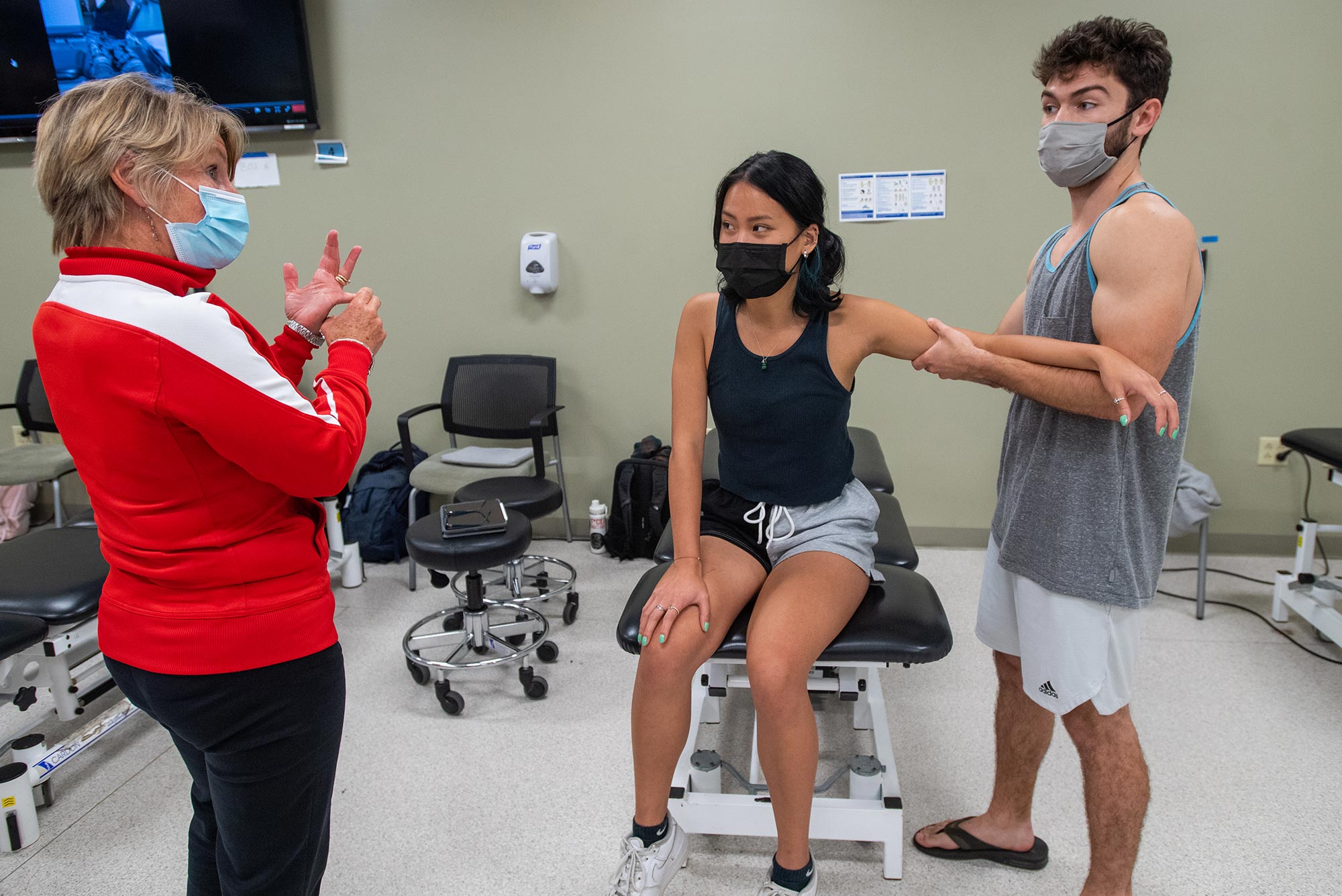 Photo of SAR clinical associate professor Lee Marinko, at left, in a red and white sweater, and Nikita Chou (SAR’24), center, sits on a physical therapy table, and Dominick Farino (SAR’24), who raises Chou’s arm. Each person wears a face mask. Chou and Farino look towards Marinko as she explains something.