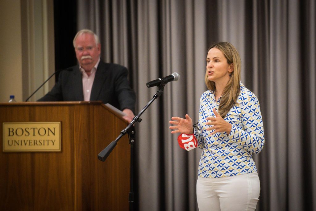 Photo of Dr. Judy Platt, in white pants and a textured blouse, speaking into a microphone on stage, as she discusses COVID protocols for the fall.