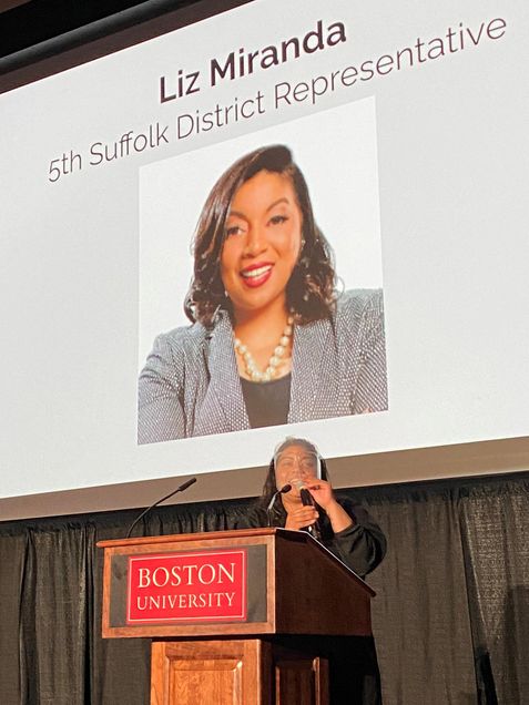 Photo of State Rep Liz Miranda addressing 2021's 400+ FYSOP students on Thursday August 26. She wears a black outfit and a face shield, and gestures as she speaks in a mic she holds at a Boston University podium. Projected behind her is her headshot and name, “Liz Miranda, 5th Suffolk District Representative.”