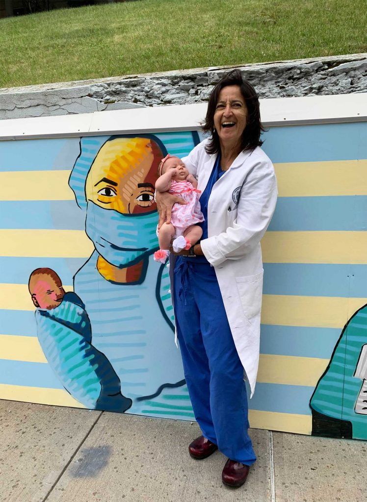 Photo of St. Elizabeth’s obstetrics and gynecology chair Isabelle Morais and infant Paisley Thomas pose in front of their portrait that Sam painted. Morais wears navy blue scrubs and a white coat, and the baby is dressed in pink. In the portrait, Morais has a face mask on and swaddles the baby in a blue towel.