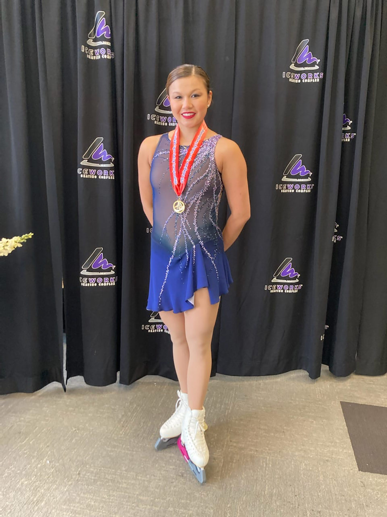 Photo of Lily Rauh in her blue and silver figure skating competition outfit with skates and her first place medal on. She smiles as he stands in front of a black curtain.