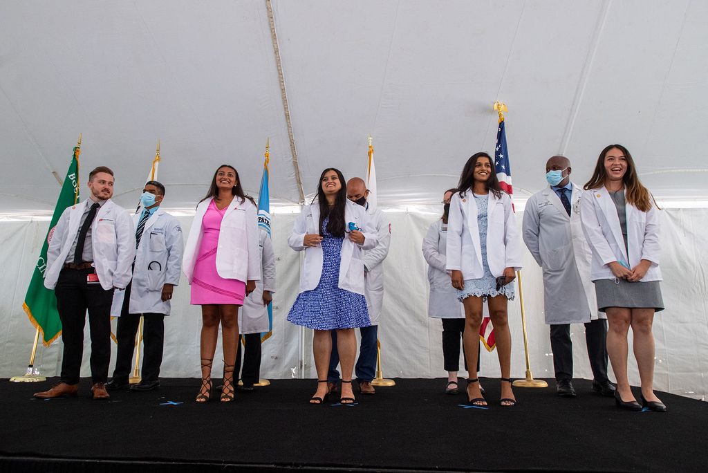 Photo of BU School of Medicine’s White Coat Ceremony on the Talbot Green August 2. In the front row, Austen Mauch, from left, Nisha Mathur, Saaz Mantri, Avni Madhani, and Kendra Lujan stand and receive their white coats. Behind them, staff and/or others receiving their coats are seen. They stand underneath a white tent on a stage on Talbot green.