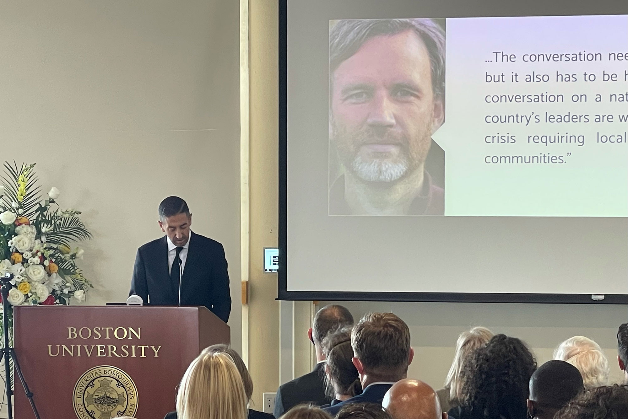 Photo of School of Public Health Dean Sandro Galea at a crimson Boston University podium, looking down and speaking into a mic, at a tearful memorial for David Jones. On a screen behind him, a photo of David Jones is seen—he’s a middle-aged man, smiling with salt-and-pepper hair and beard. A quote of his is next to his portrait but it’s cut off. The backs of peoples heads who are seating are seen in the foreground.