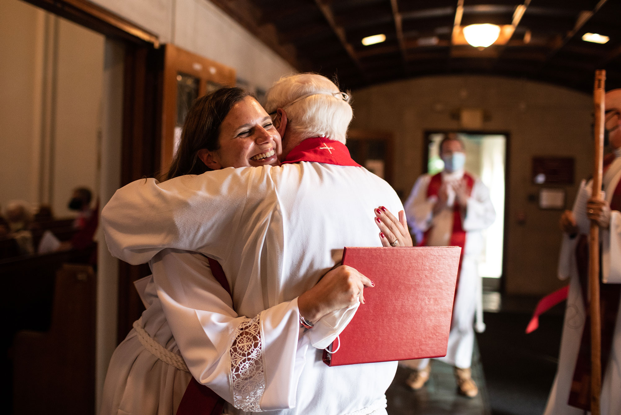 Photo of Dr. Jessica Chicka hugging her father, Raymond C. Hittinger, a retired Lutheran pastor, after her ordination at Marsh Chapel, Sunday, September 12, 2021. Both wear long white robes. Chicka smiles wide and holds a red binder; the back of Hittinger’s head is seen.