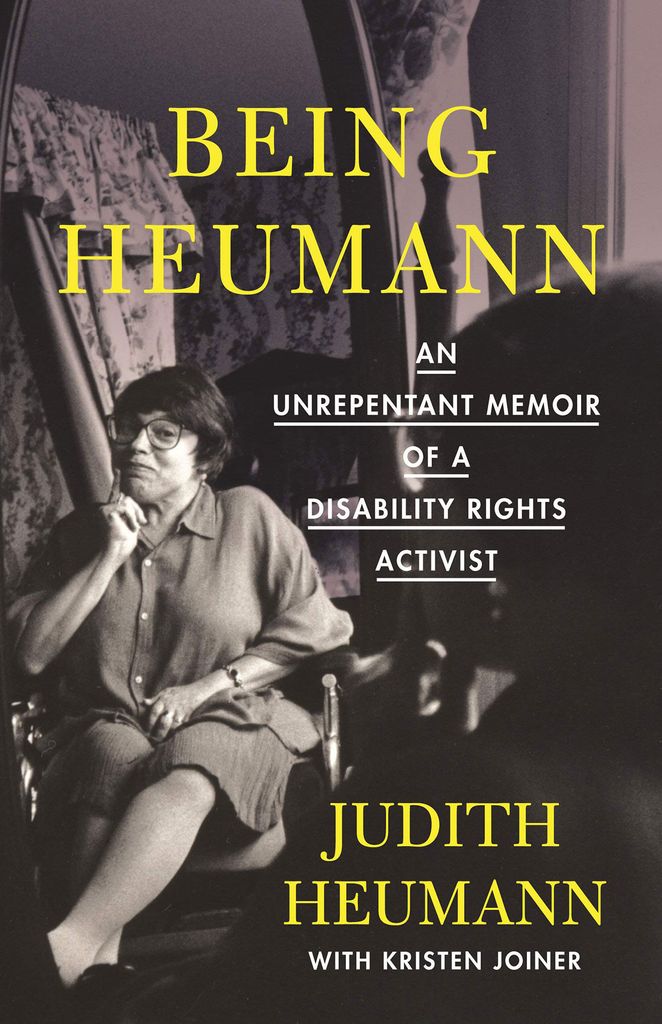 Book cover for "Being Heumann: An Unrepentant Memoir of a Disability Rights Activist". The title is in bright yellow font. The cover is a black and white photo of Judith sitting in her wheelchair with a pant suit on and her hand on her face. She smiles and looks into a mirror. At the bottom, “Judith Heumann with Kristen Joiner” is written.