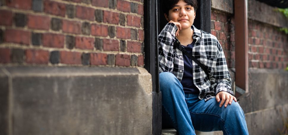 Photo of Ash Vasquez (CFA’25), sitting on a step between two brick walls. His hand is on his face, and he wears a plaid shirt and jeans.