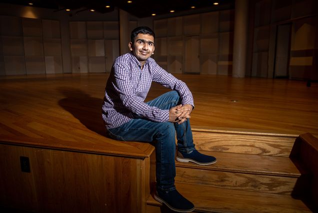 Photo of Faizaan Firoz (Pardee’25) sitting on the stairs leading up to a stage with a checkered button down and jeans on. He smiles and the dramatic lighting casts a long shadow on the stage behind him.