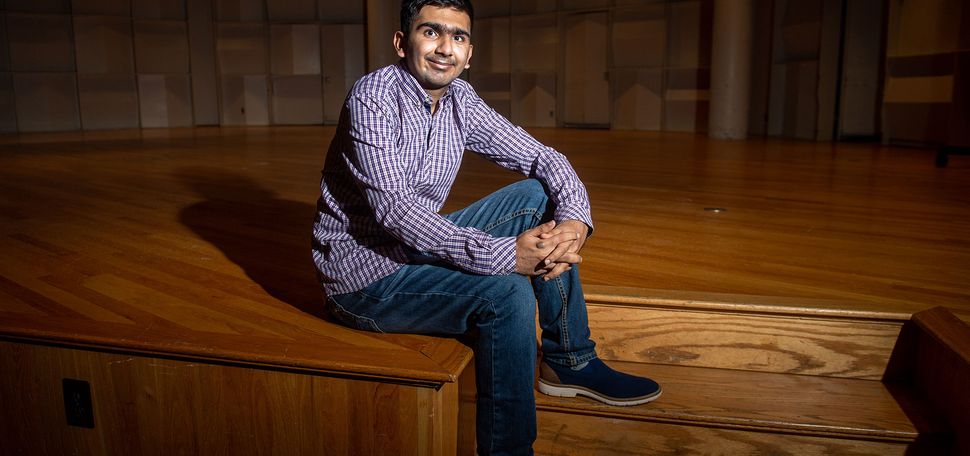 Photo of Faizaan Firoz (Pardee’25) sitting on the stairs leading up to a stage with a checkered button down and jeans on. He smiles and the dramatic lighting casts a long shadow on the stage behind him.