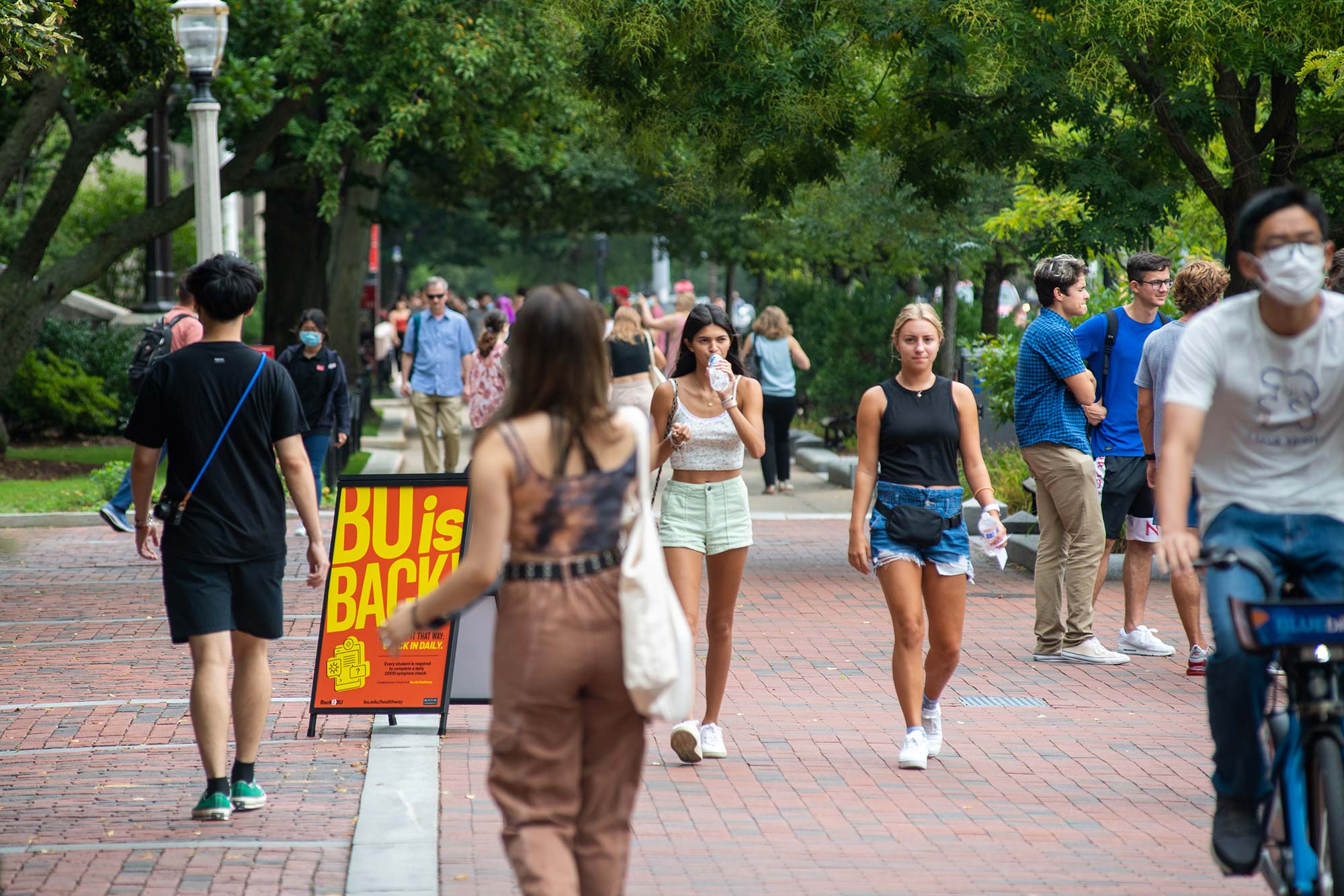 Photo of students walking down Comm Ave past an orange and yellow sign that says "BU is Back". Students are dressed in summer attire and the trees along the street are green.