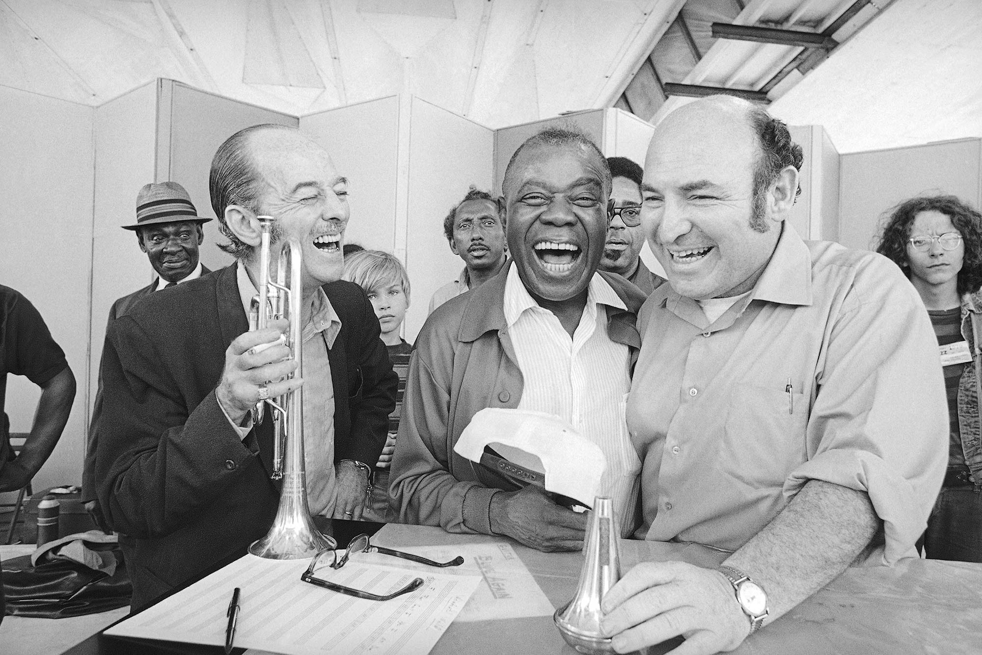 Black and white photo of Louis Armstrong, center, joins in his own joke over an old jazz story with trumpeter Bobby Hackett and Newport Jazz Festival impresario George Wein, at left, at Newport, Rhode Island on July 10, 1970. Hackett holds a trumpet, Armstrong is mid-laugh, and all the men smile.