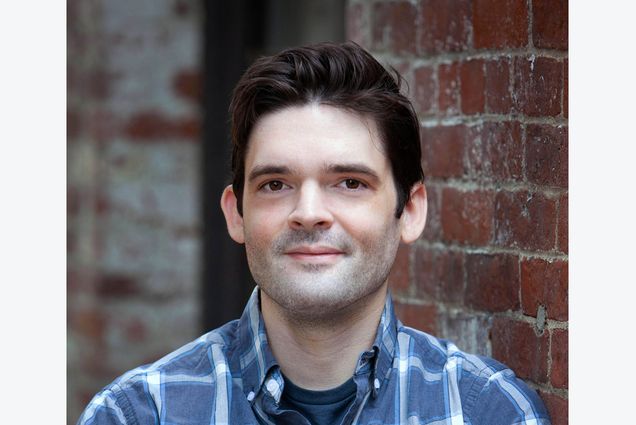 Headshot of Paul Coleman (COM’21) wearing a blue and white plaid shirt with his arms across his chest. He smiles slightly and leans against a brick wall. He has dark brown hair.