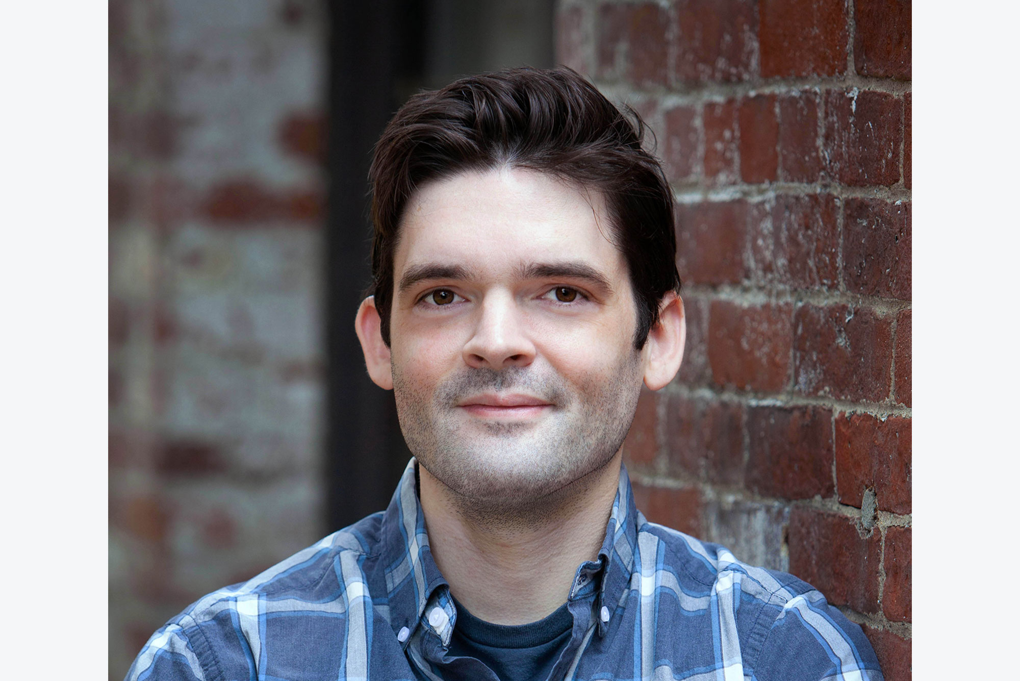 Headshot of Paul Coleman (COM’21) wearing a blue and white plaid shirt with his arms across his chest. He smiles slightly and leans against a brick wall. He has dark brown hair.