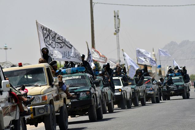 Photo of Taliban fighters atop vehicles with white Taliban flags with black characters, parading along a road to celebrate after the US pulled all its troops out of Afghanistan, in Kandahar on September 1, 2021 following the Talibans military takeover of the country. The cars are mostly dark colored trucks and the fighters all appear to be men.