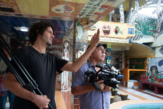 Photo of Director Ken Schneider (left) working with director of photography Roberto Chile at the home of Cuban artist Fuster for the film, Los Hermanos/The Brothers, which is one of the nine films screening during BLIFF. Schneider has brown curly hair and a gotee and holds a tripod. Chile holds a video camera and both men look off to their left. They are in a building covered by mosaic designs.