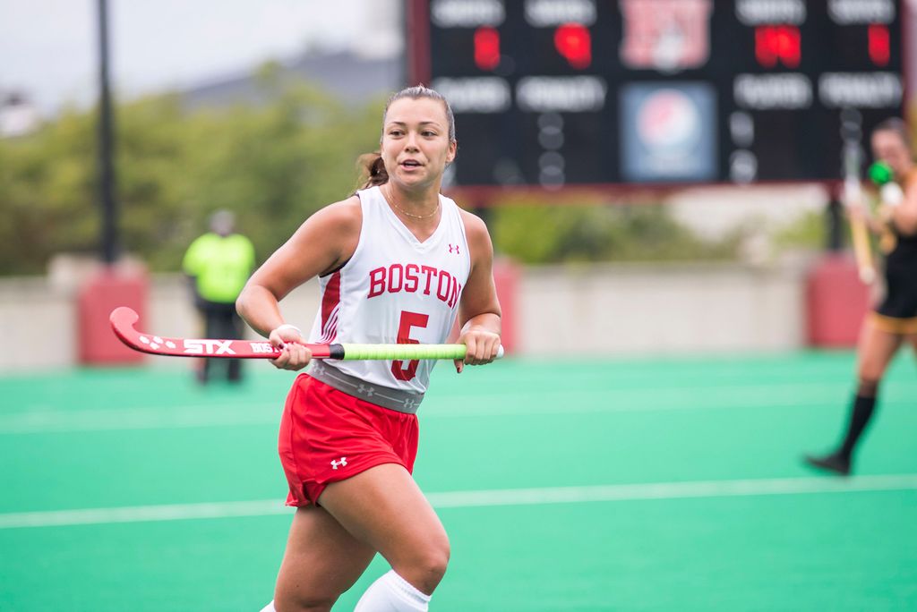 Photo of Miya Denison, wearing a white and red field hockey jersey with the number 5, and holding a red and green hockey stick as she runs across the field.