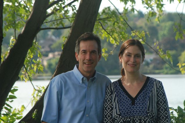 Photo of Andrew Budson in a blue button down shirt and Dr. Maureen O’Connor, in a patterned blue, white and black blouse, posing for a headshot in front of some trees and a lake.