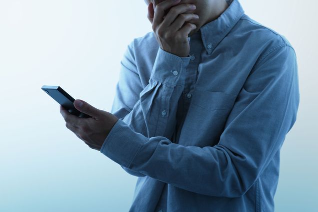 Photo of a young person in a blue button down shirt holding their phone and putting their hand over their mouth in shock.