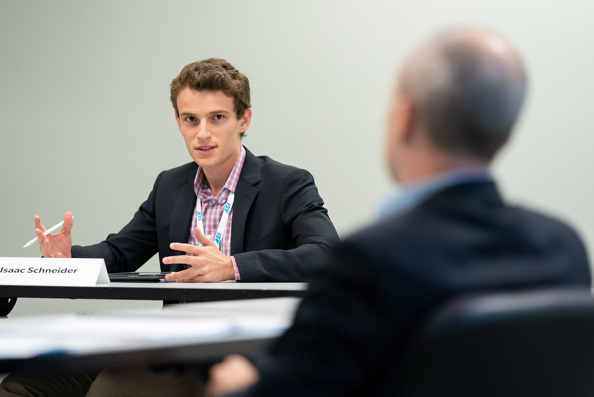 Photo of Zak Schneider, in a black suit jacket and red and white checkered shirt behind a desk with a name tag on it, talking to a person who is blurred in the right foreground, who is identified as AEI Scholar Yuval Levin.