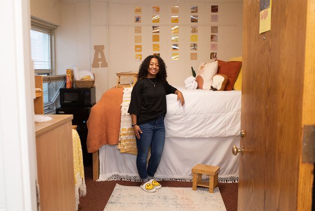 Photo of Anea Harris (Wheelock'24), in a black shirt, blue jeans, and slippers with smilie faces on them, resting her hand on her lofted bed in her dorm room. The walls are decorated with photos and the bed has pillows in the shape of a knot and a piece of toast.